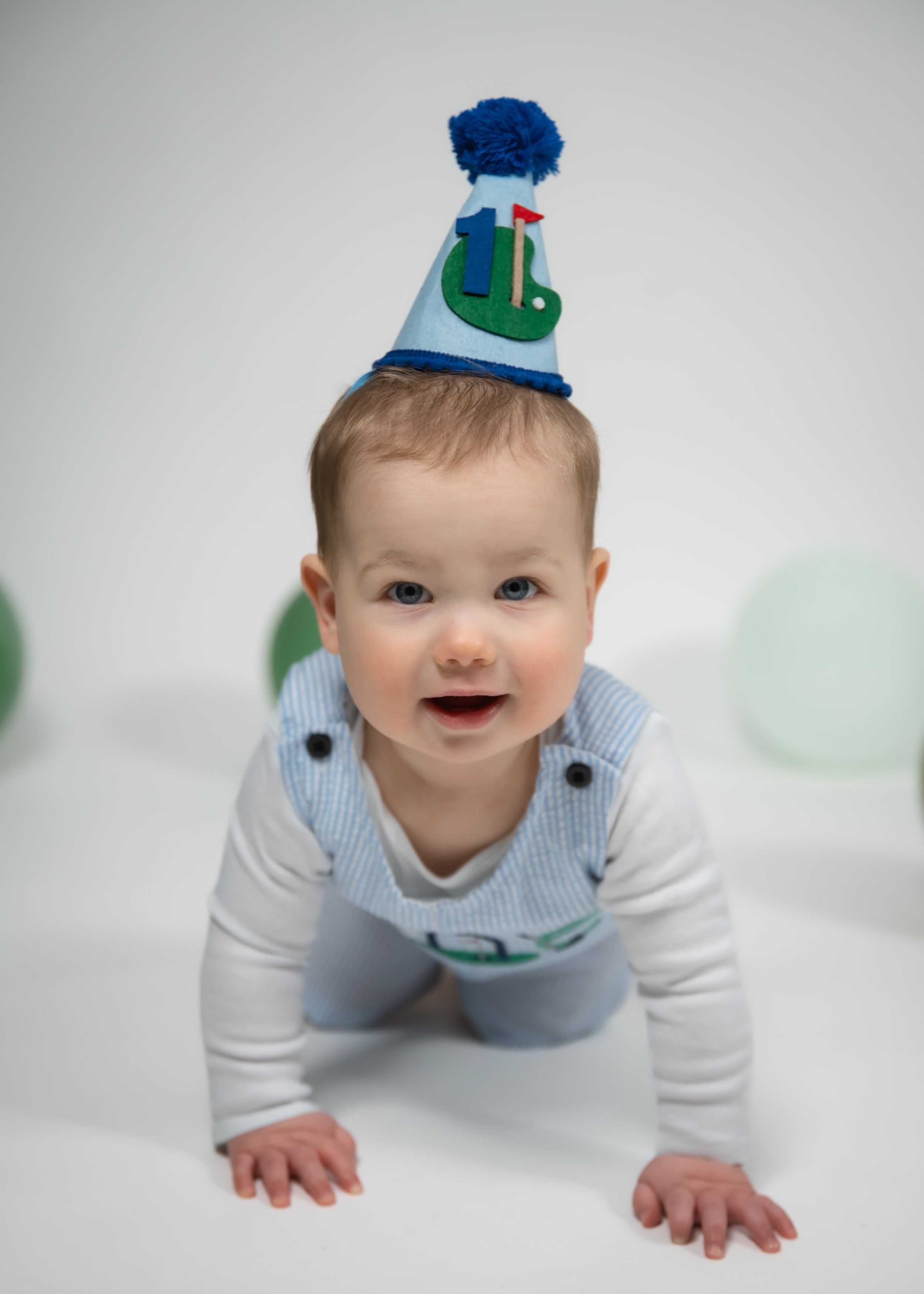 A smiling baby wearing a golf-themed birthday party hat crawls on a white background in a blue outfit.