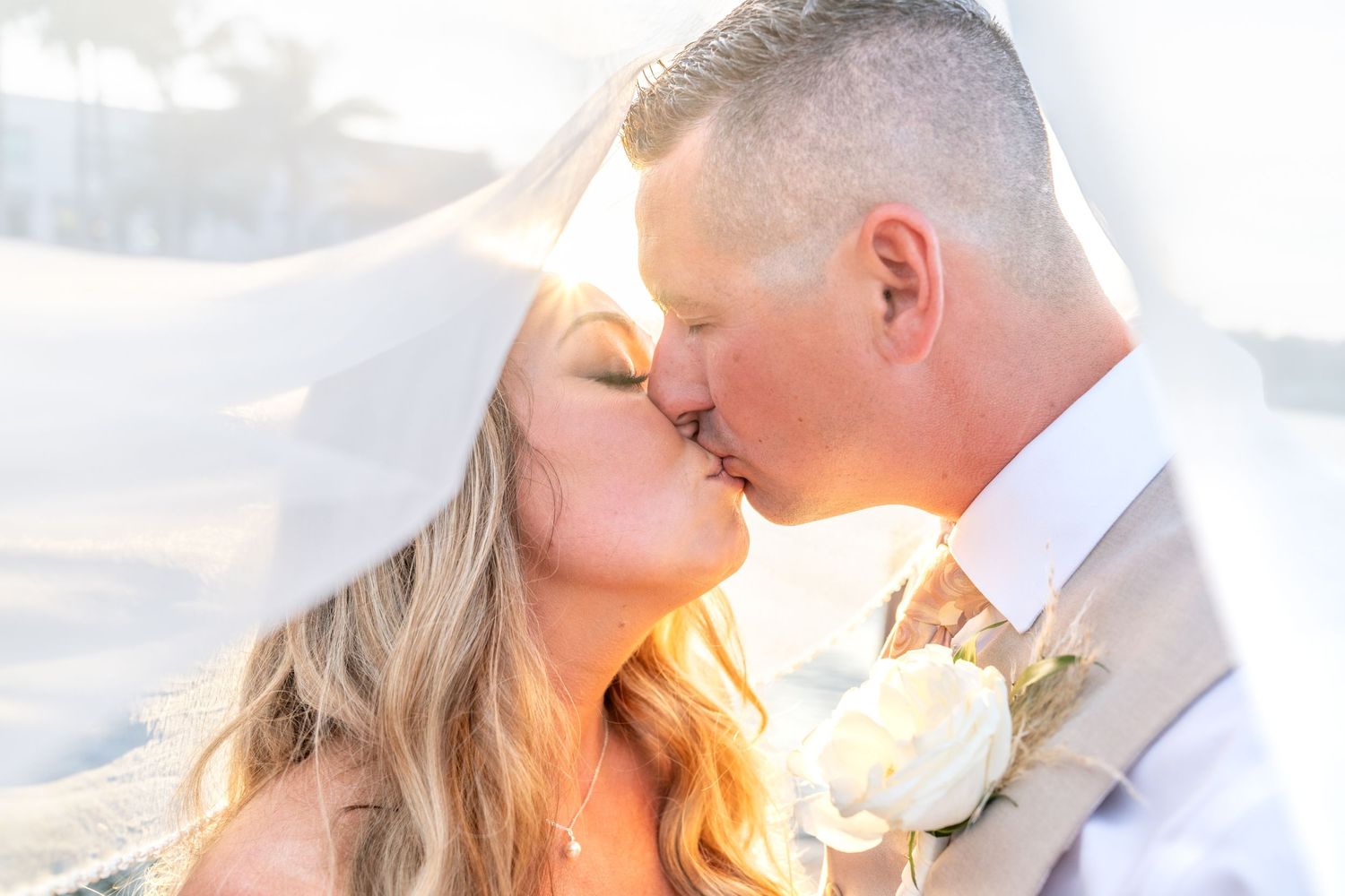 Bride and groom share a romantic kiss under a flowing wedding veil with warm sunlight glowing behind them.