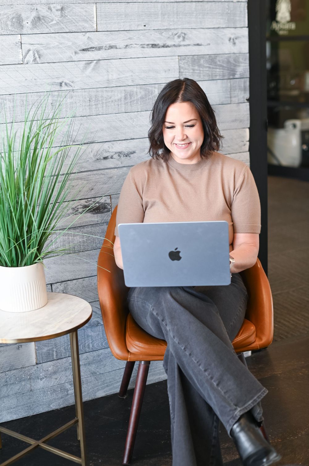 Woman smiling while using a MacBook laptop, seated in a brown leather chair next to a potted plant.