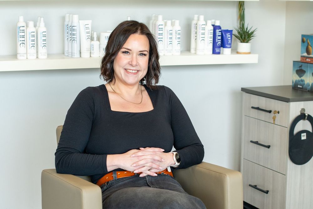 Woman smiling in a modern salon chair with hair care products displayed on shelves in the background.