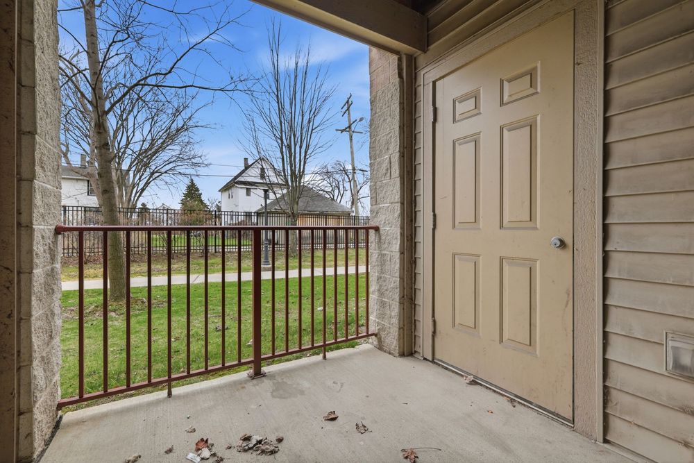 Covered front porch with beige door, metal railing, concrete floor, and a view of a green lawn and neighboring homes. Detroit MI