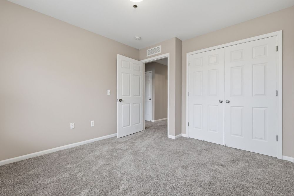 Spacious bedroom with gray carpet, white closet doors, and neutral beige walls in a modern apartment. Detroit MI