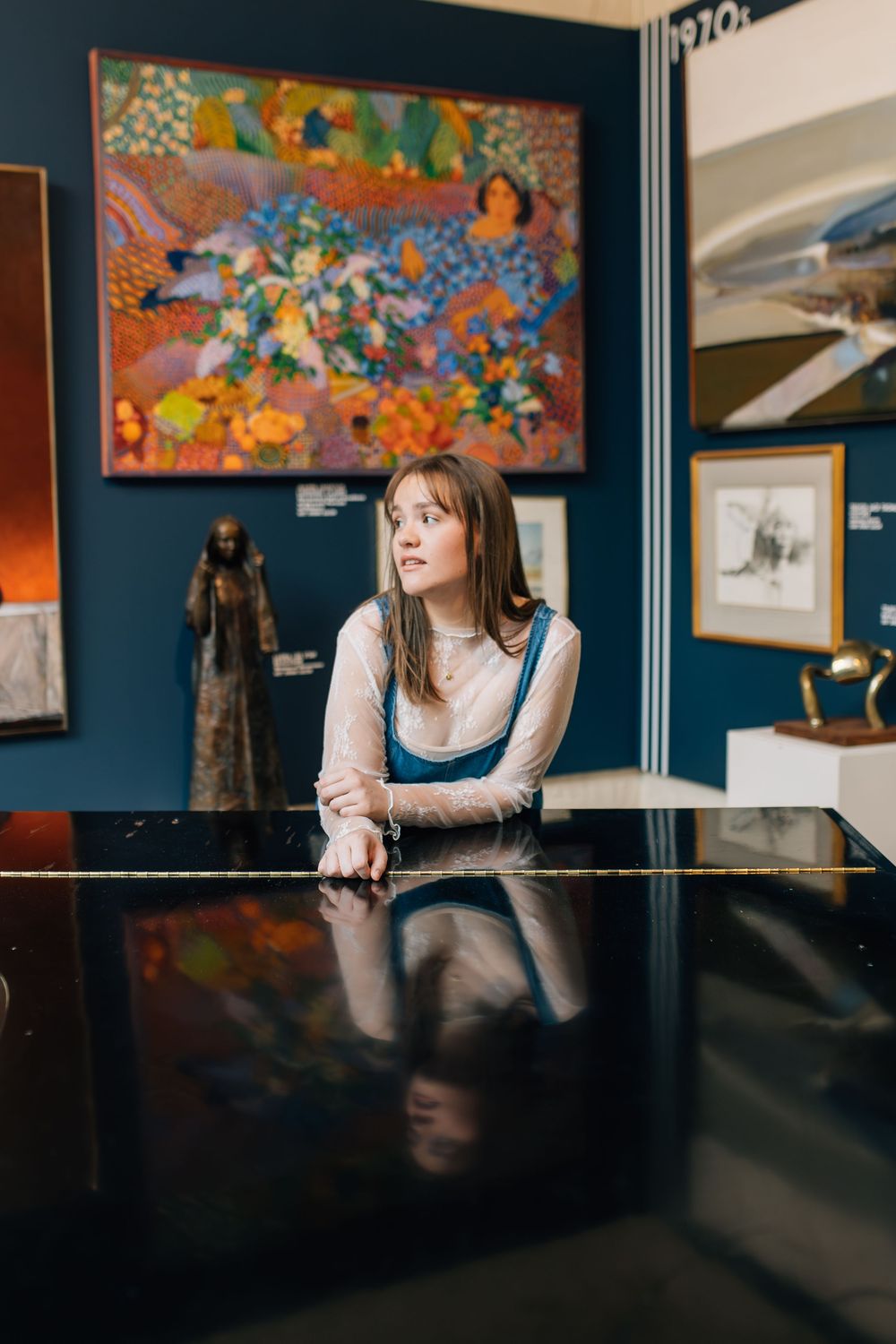 Young woman in denim overalls leans on a piano in an art gallery with colorful paintings on display.