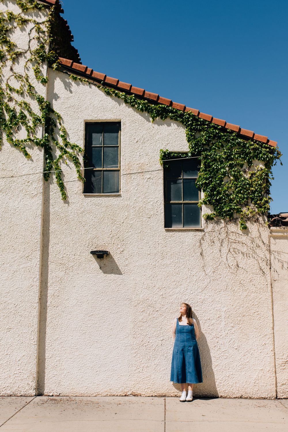 Young woman in denim dress leaning against ivy-covered white stucco building under bright blue sky.