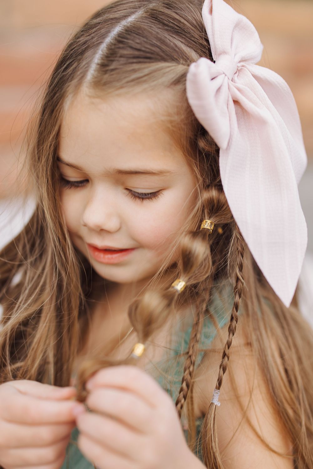 Close-up of young girl with long hair, pink bow, and braids looking down thoughtfully.