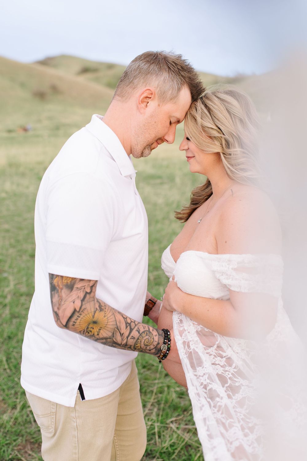Couple touching foreheads tenderly in grassy field, man with tattoo sleeve in white shirt.