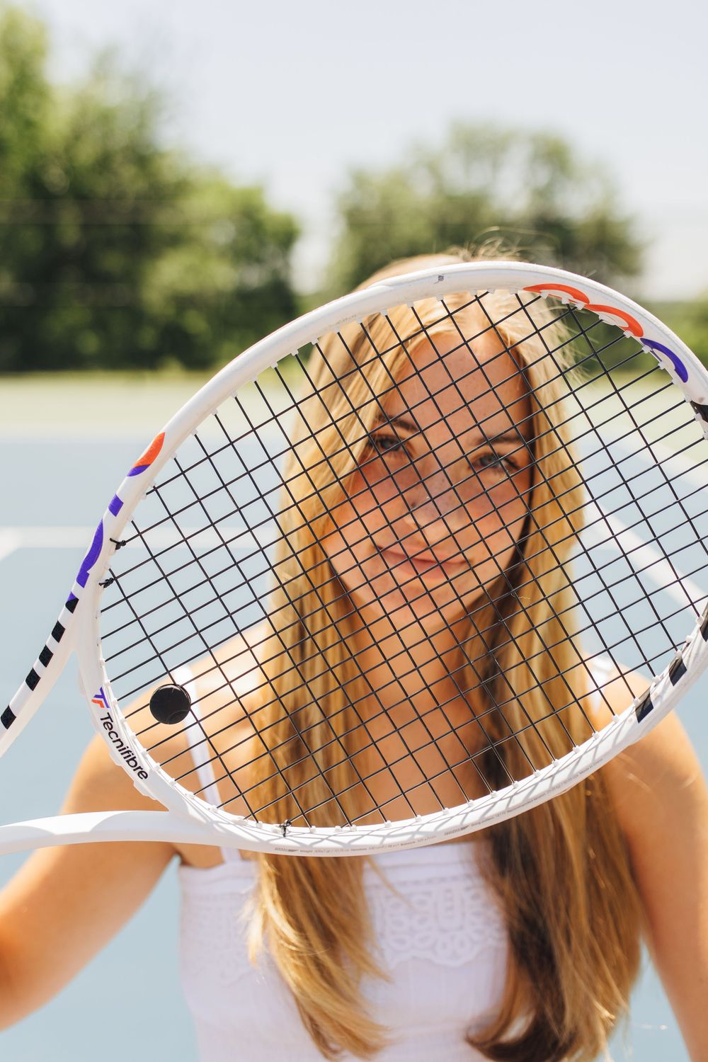 Smiling blonde woman peers through tennis racket strings on a sunny outdoor court.