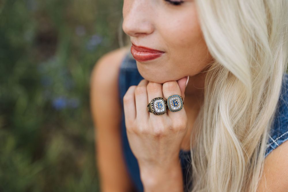 Close-up of blonde woman wearing two sparkling championship rings against blurred floral background.