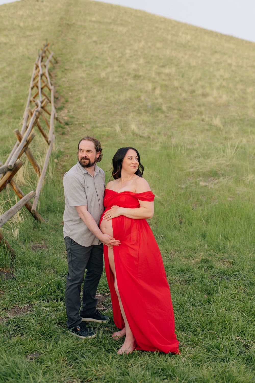 Expecting couple stands on green hillside, woman in red maternity gown, partner embracing her.