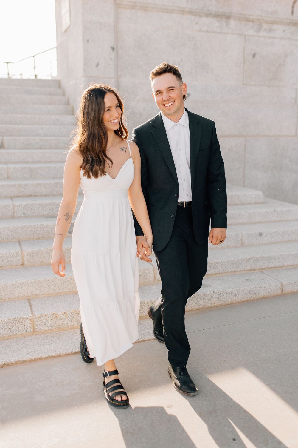 Happy couple holding hands walking down sunlit steps, woman in white maxi dress and black sandals.