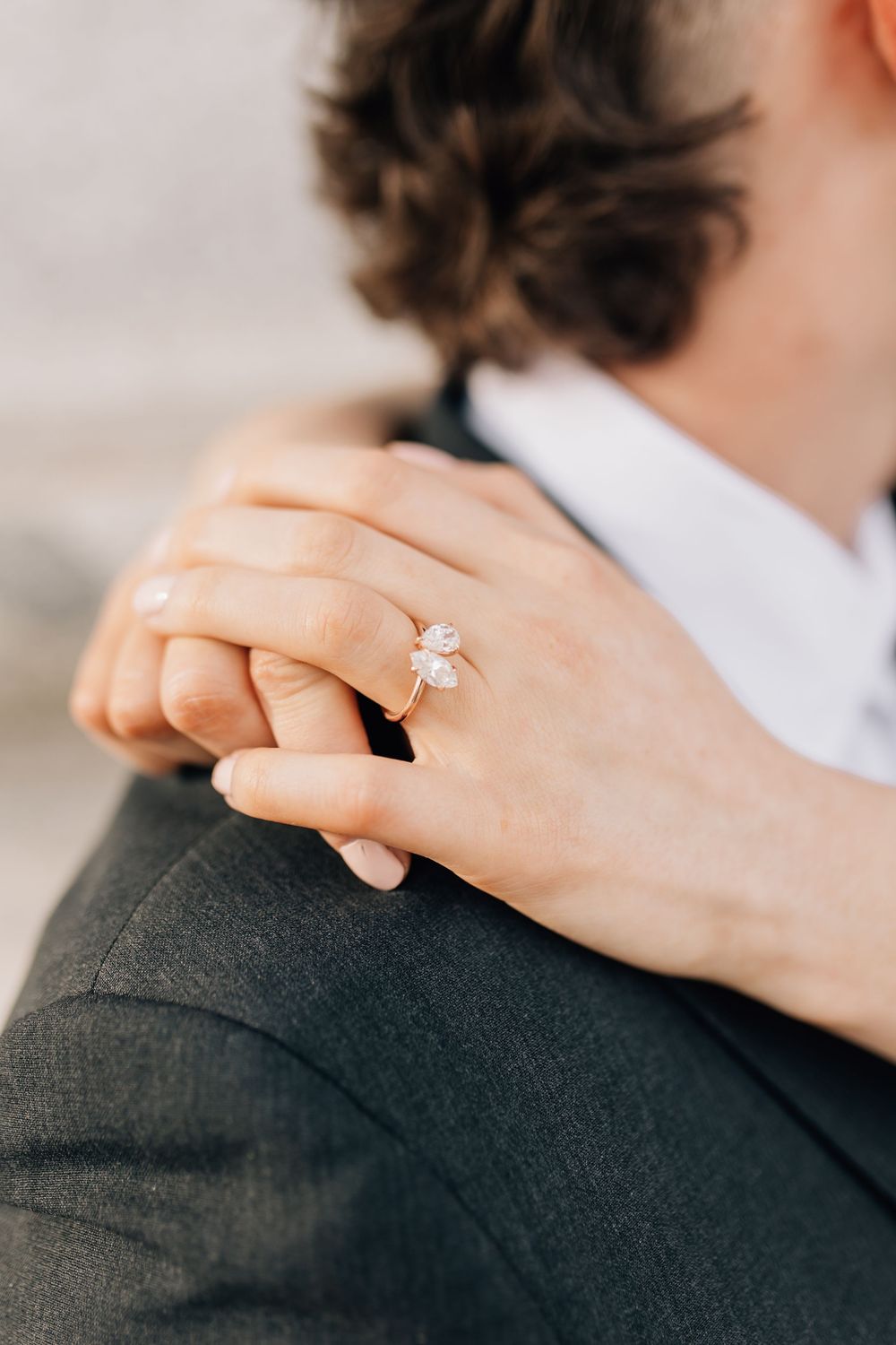 Close-up of a womans hand with elegant diamond engagement ring resting on mans dark suit shoulder.