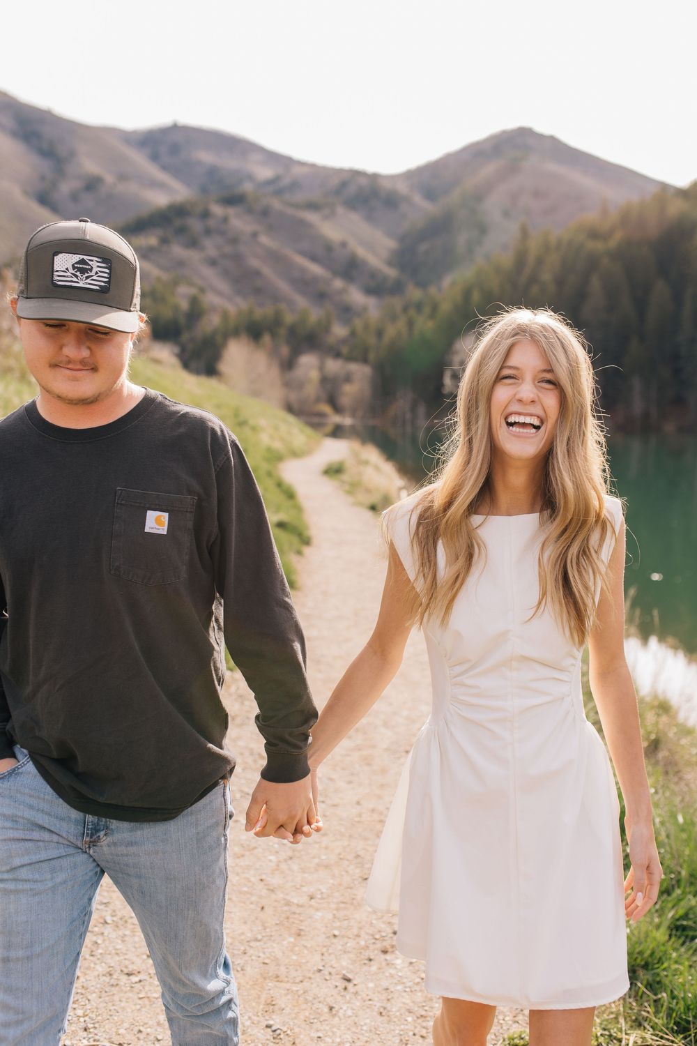 Happy couple holds hands walking on scenic mountain path beside turquoise lake on sunny day.
