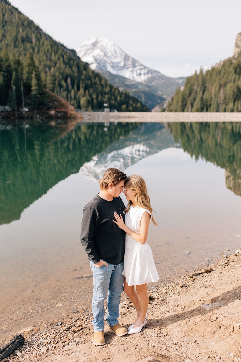 Romantic couple nearly kissing beside a calm reflective alpine lake with snow-capped mountain backdrop.