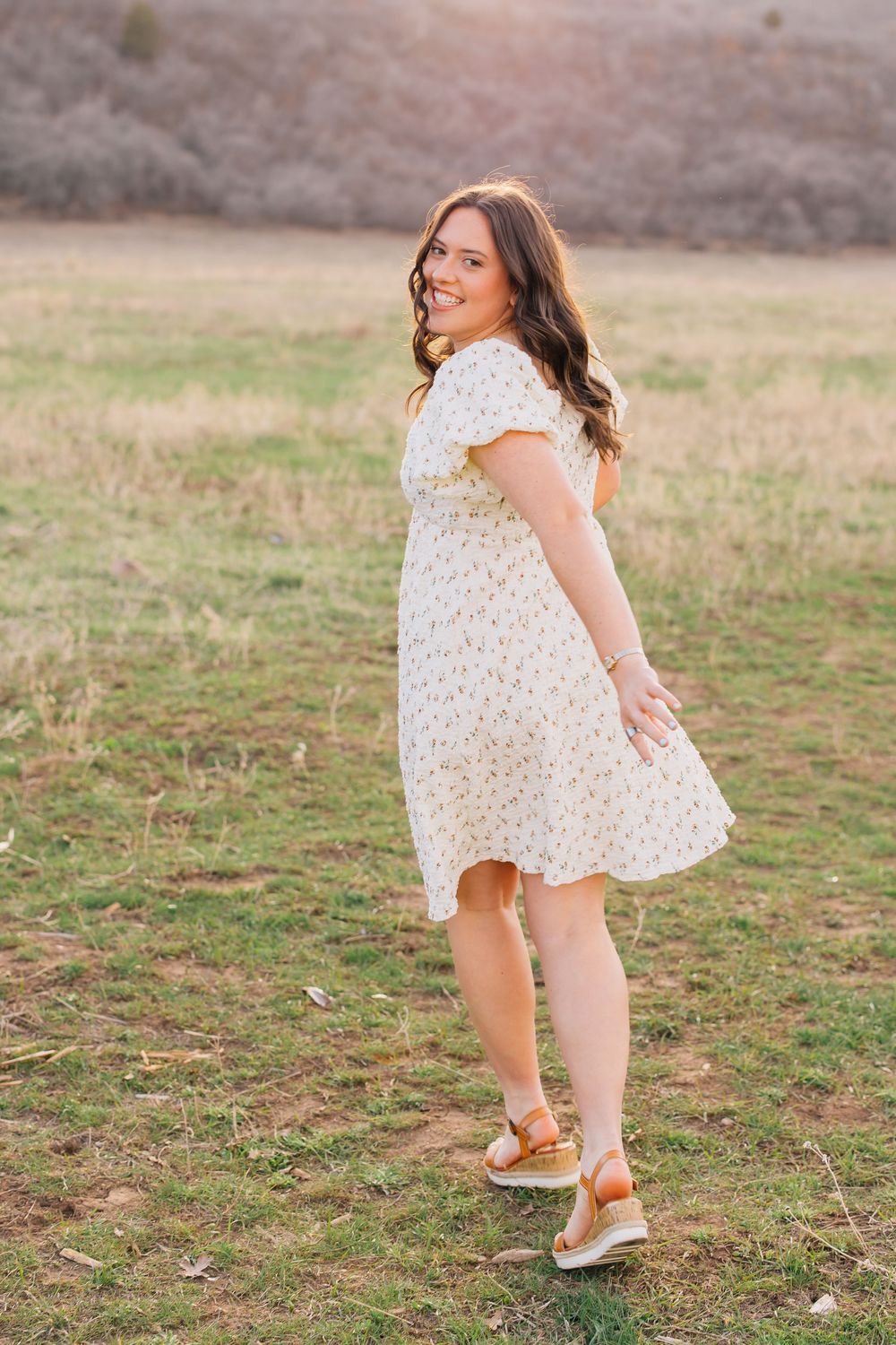 Smiling brunette woman in floral dress twirling in golden hour sunlit open field.