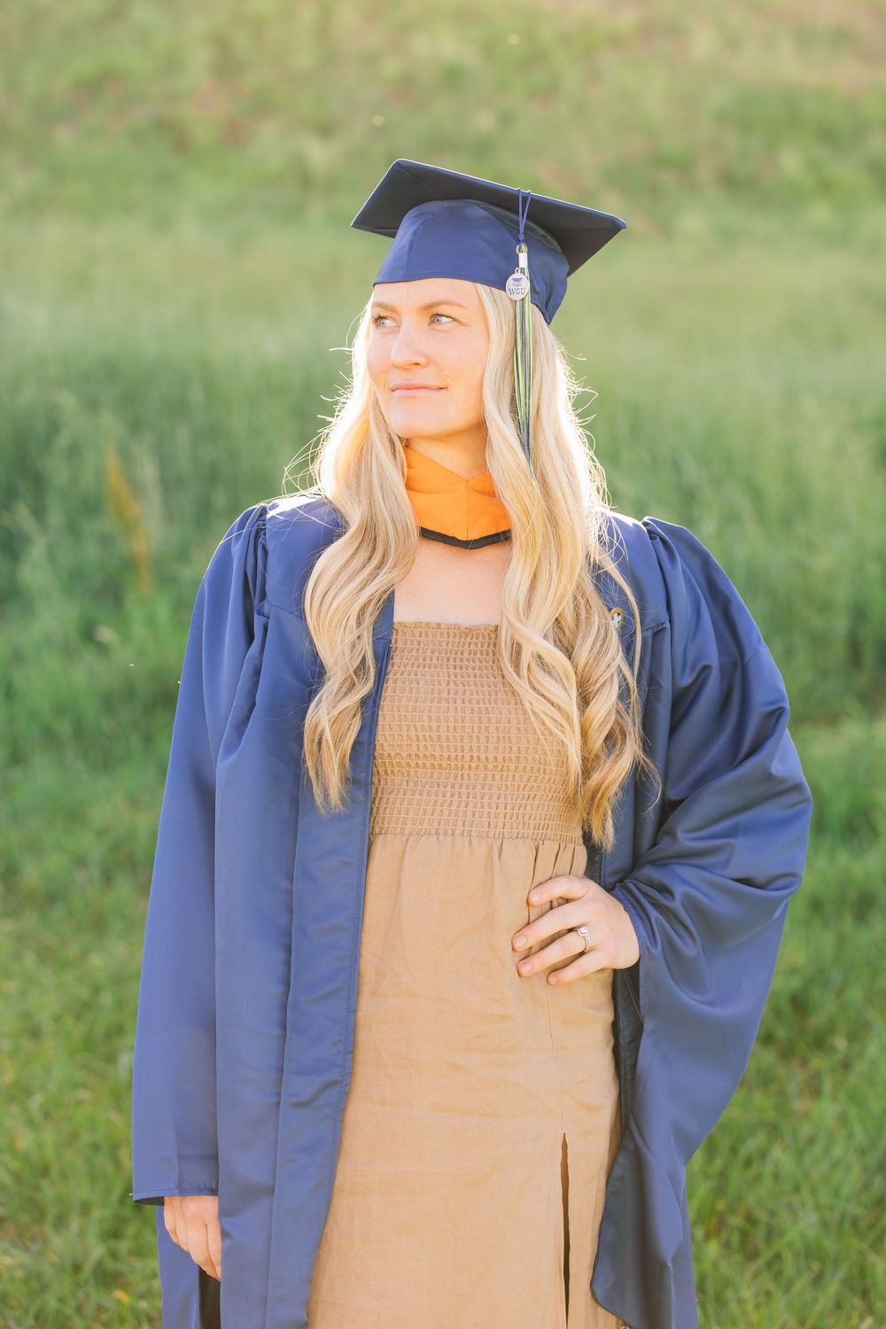 Blonde woman in blue graduation cap and gown posing in sunlit green field.