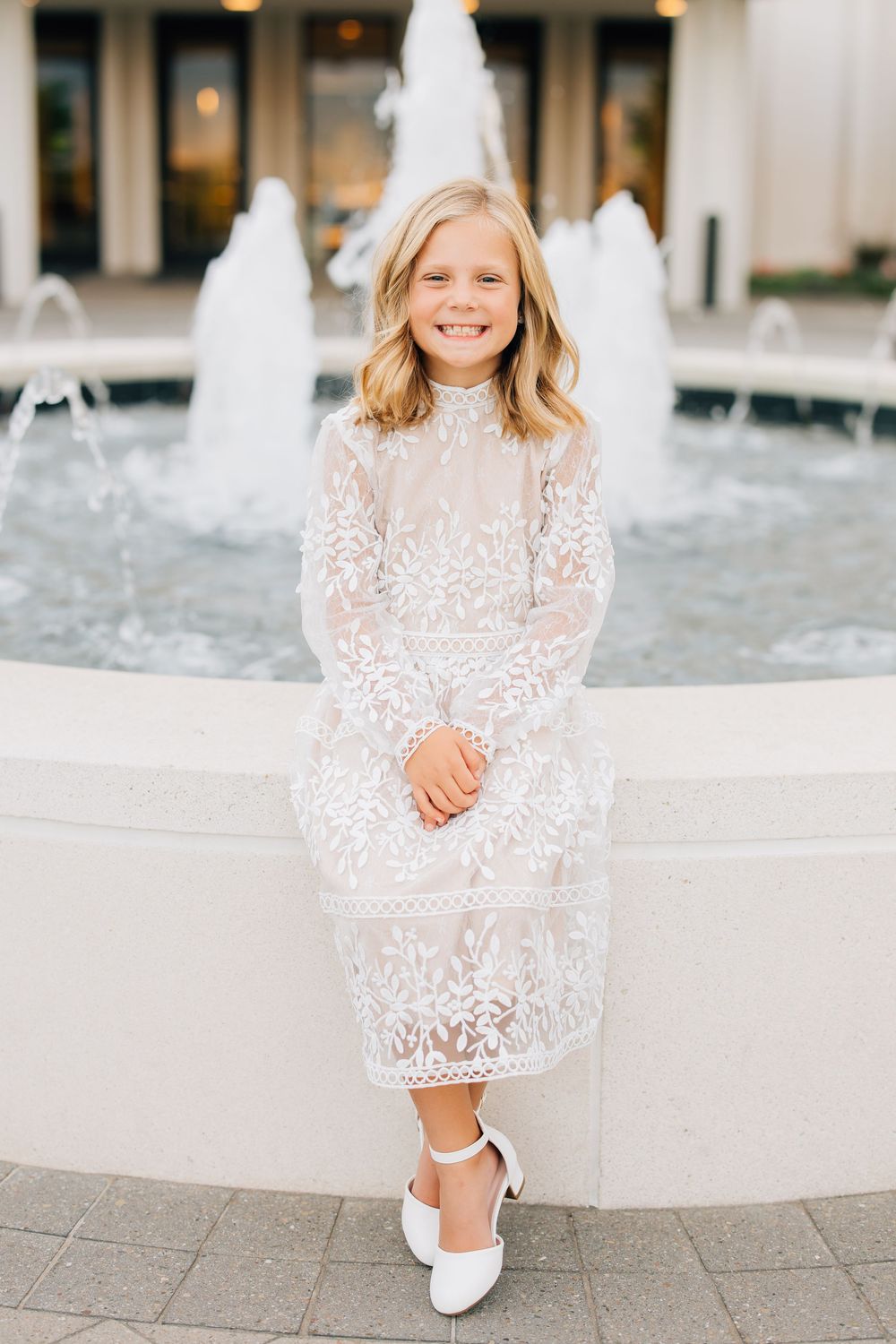 Smiling young girl in white lace dress stands before a fountain in elegant white Mary Jane shoes.