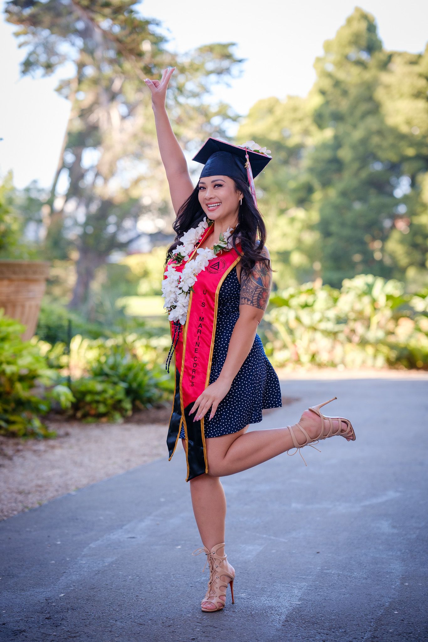 Smiling graduate in cap, gown, and floral lei poses joyfully outdoors, raising one arm and leg in celebration.
