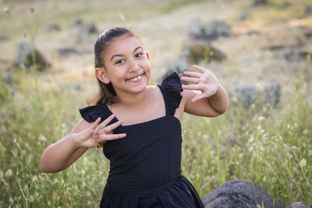 Young girl in black dress smiling and posing with hands outstretched in a grassy outdoor field.