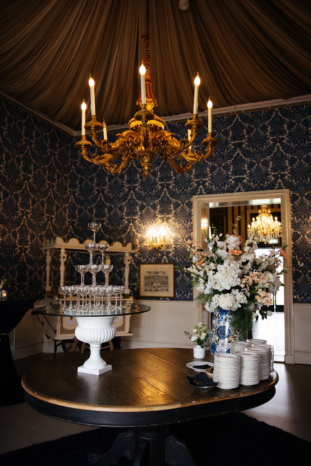 Elegant event room with gold chandelier, champagne tower, floral arrangements, and ornate blue damask wallpaper.