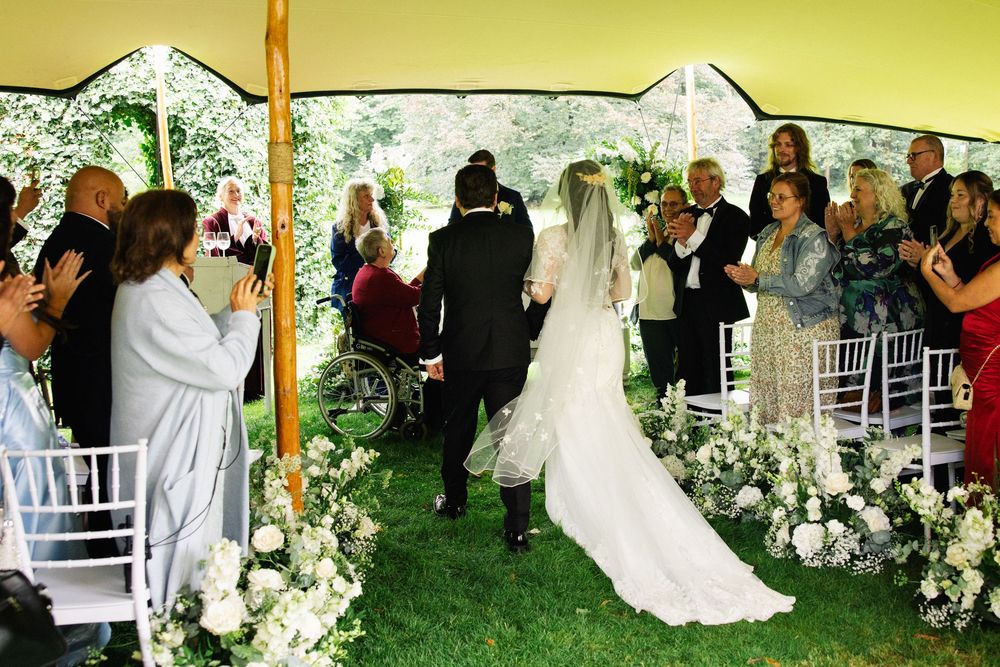 Bride walking down the aisle under a white tent surrounded by floral arrangements and guests at an outdoor wedding.