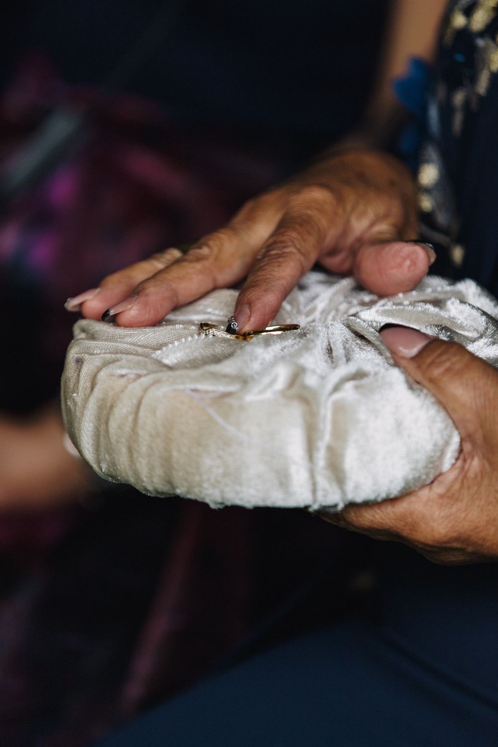 Elderly hands holding a small white velvet cushion, with the wedding rings.