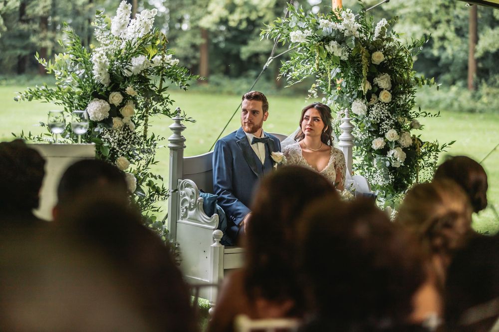 Bride and groom seated at outdoor wedding ceremony beneath floral arch with white blooms and greenery.