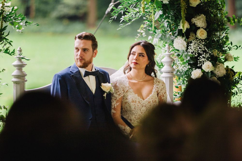 Bride and groom stand together at outdoor wedding ceremony beneath floral arch with white roses and greenery.