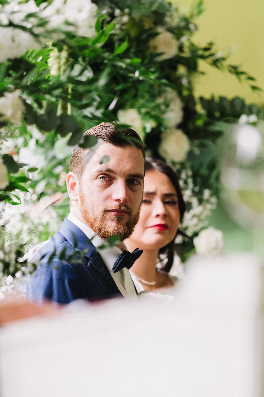 Groom in blue suit with bow tie stands beside bride at floral wedding ceremony arch with white roses.