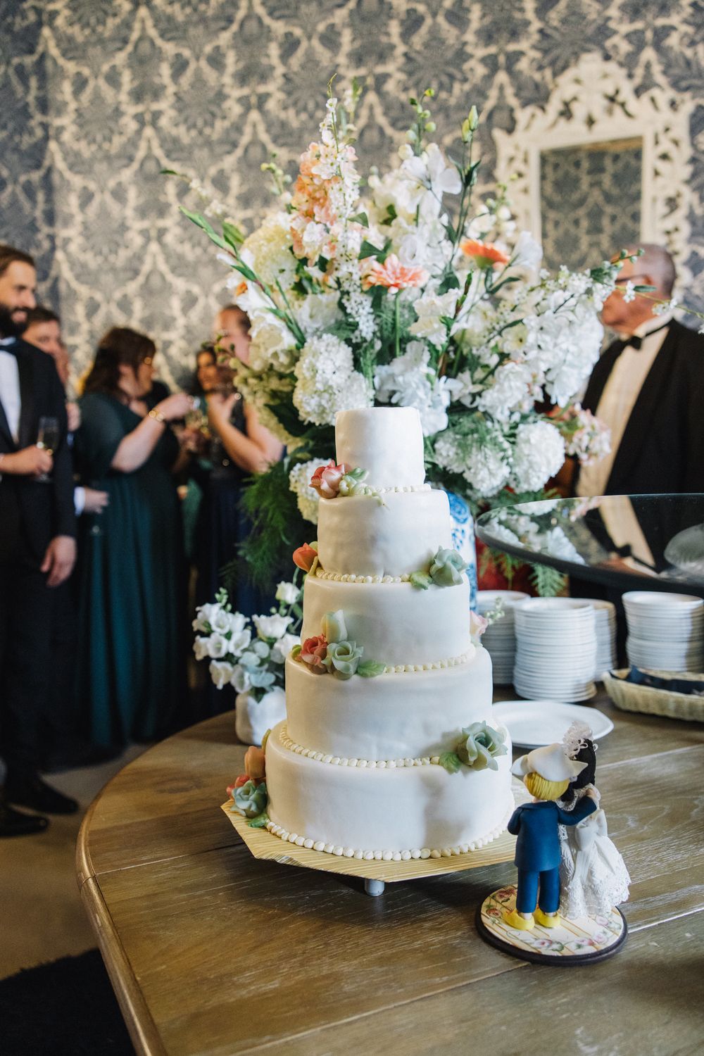 Elegant five-tier white wedding cake adorned with fresh flowers and a bride and groom topper at a wedding reception.