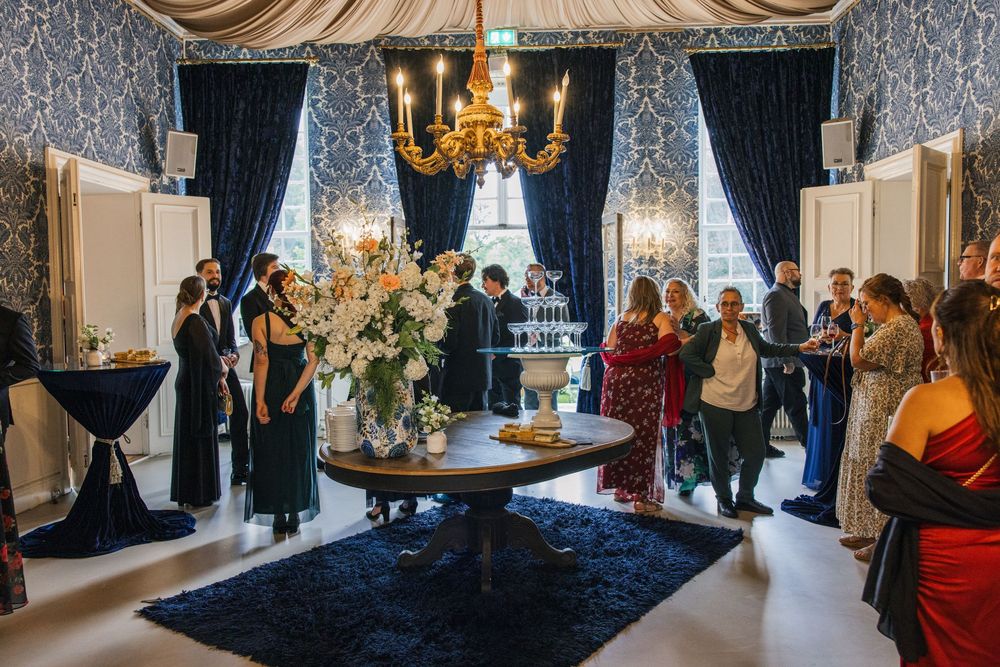 Elegant wedding reception in a grand blue-wallpapered mansion's room with chandelier, guests mingling around a floral centerpiece.