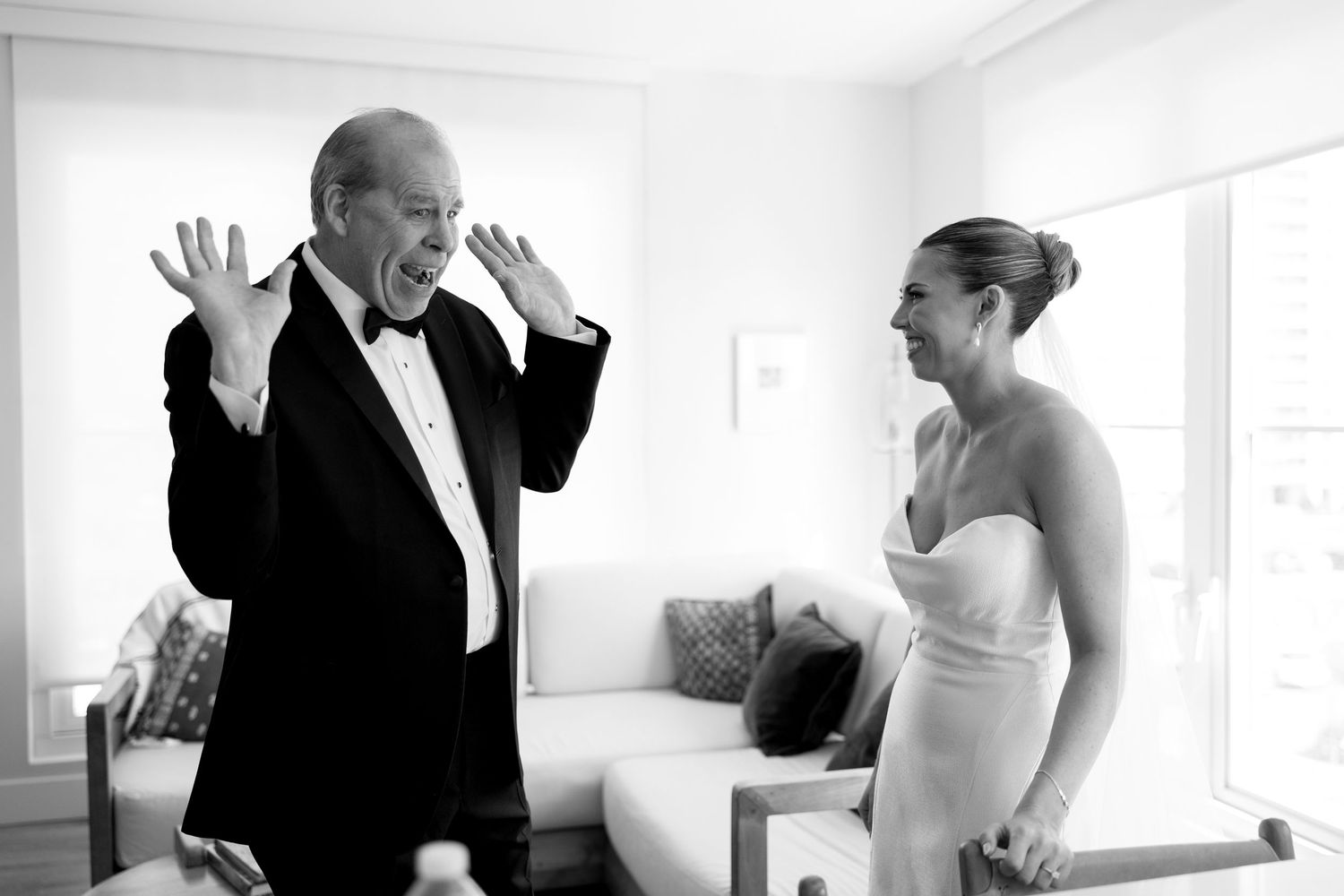 Black and white photo of a bride in a strapless gown laughing with an older man in a tuxedo.