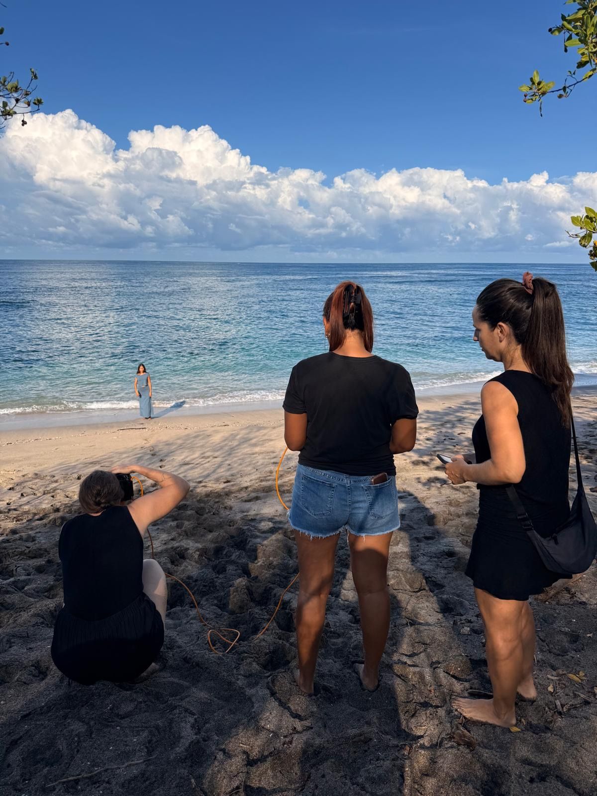 Three women on a rocky beach shore watching a fourth person pose for photos near the turquoise ocean under a blue sky.