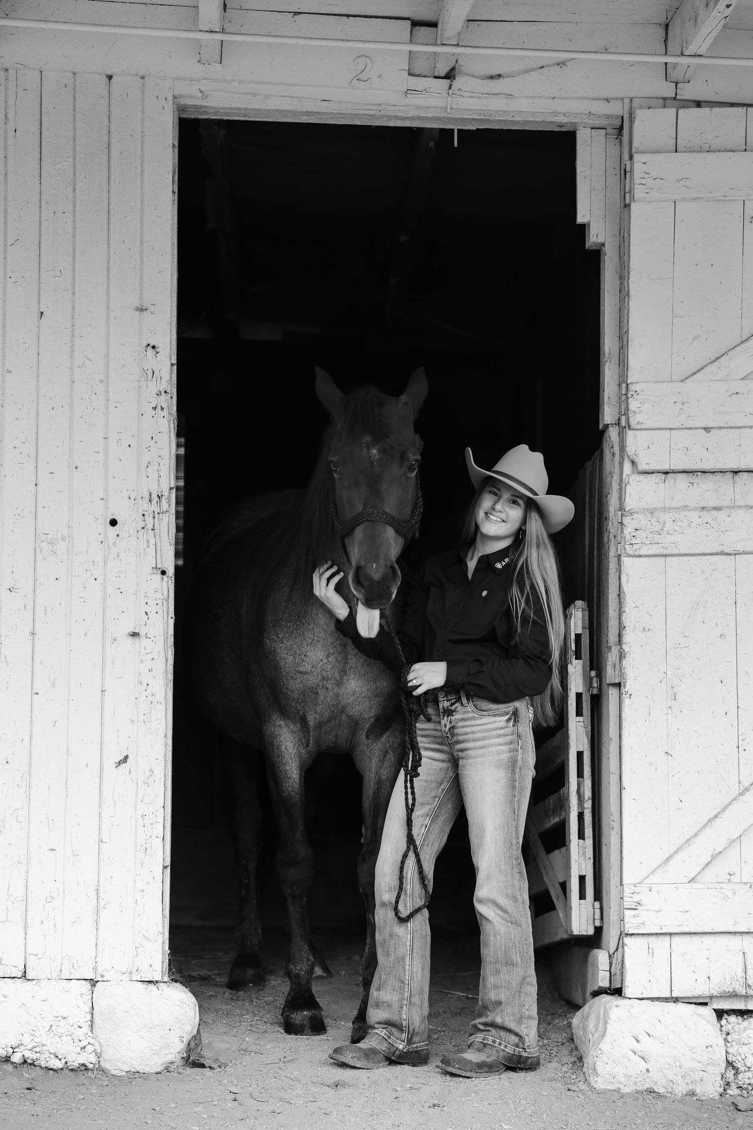 Black and white photo of a smiling woman in cowboy hat and jeans standing with a dark horse in a barn doorway.