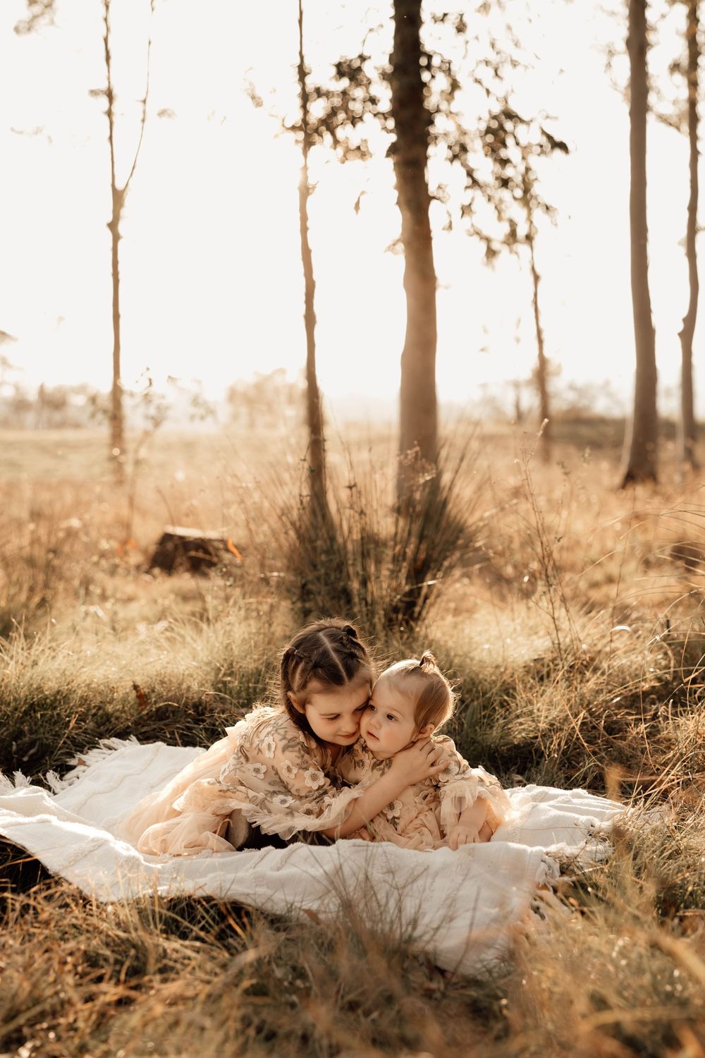Two young sisters in floral dresses share a tender embrace on a blanket in a sunlit autumn forest.