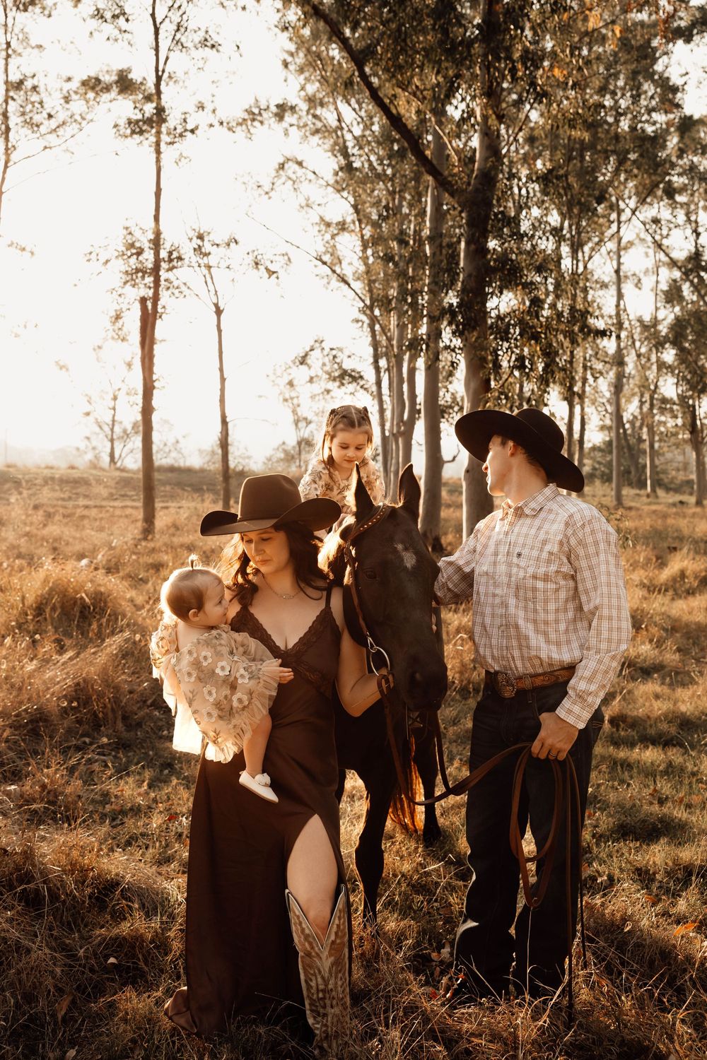 Western-style family with baby, young girl on horseback, and parents in cowboy hats at sunset.