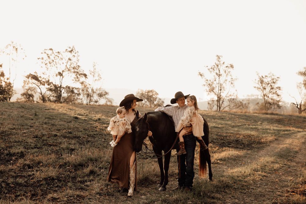 Family with two children and a dark horse on a sunlit grassy hill with trees in warm golden light.