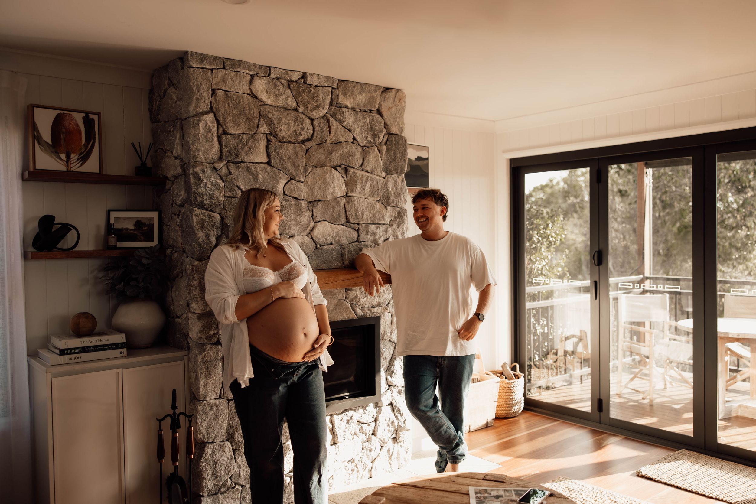 Pregnant woman and partner standing by stone fireplace in bright living room with sliding glass doors.