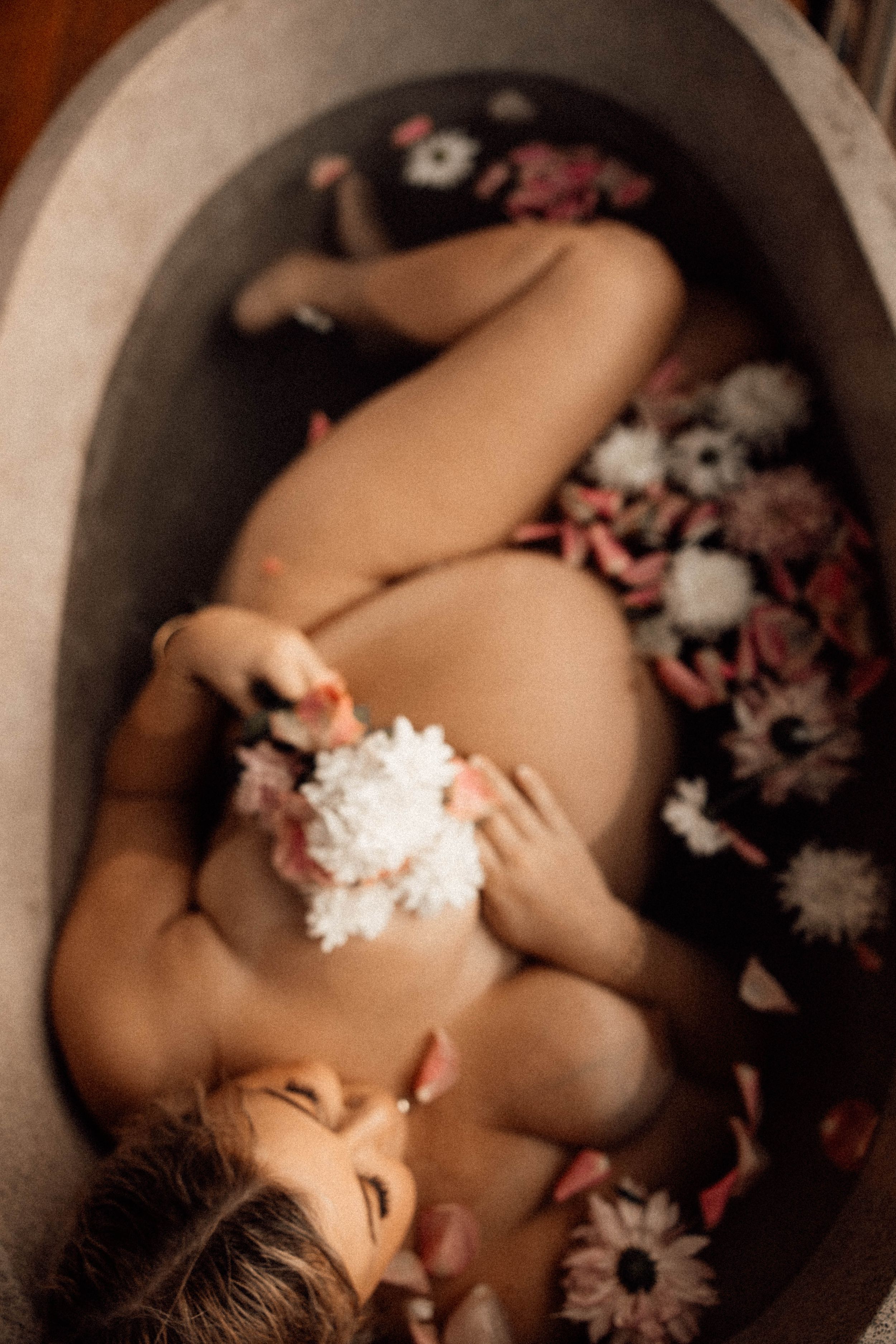 Pregnant woman relaxing in a floral milk bath, holding white flowers, captured from above in a stone tub.