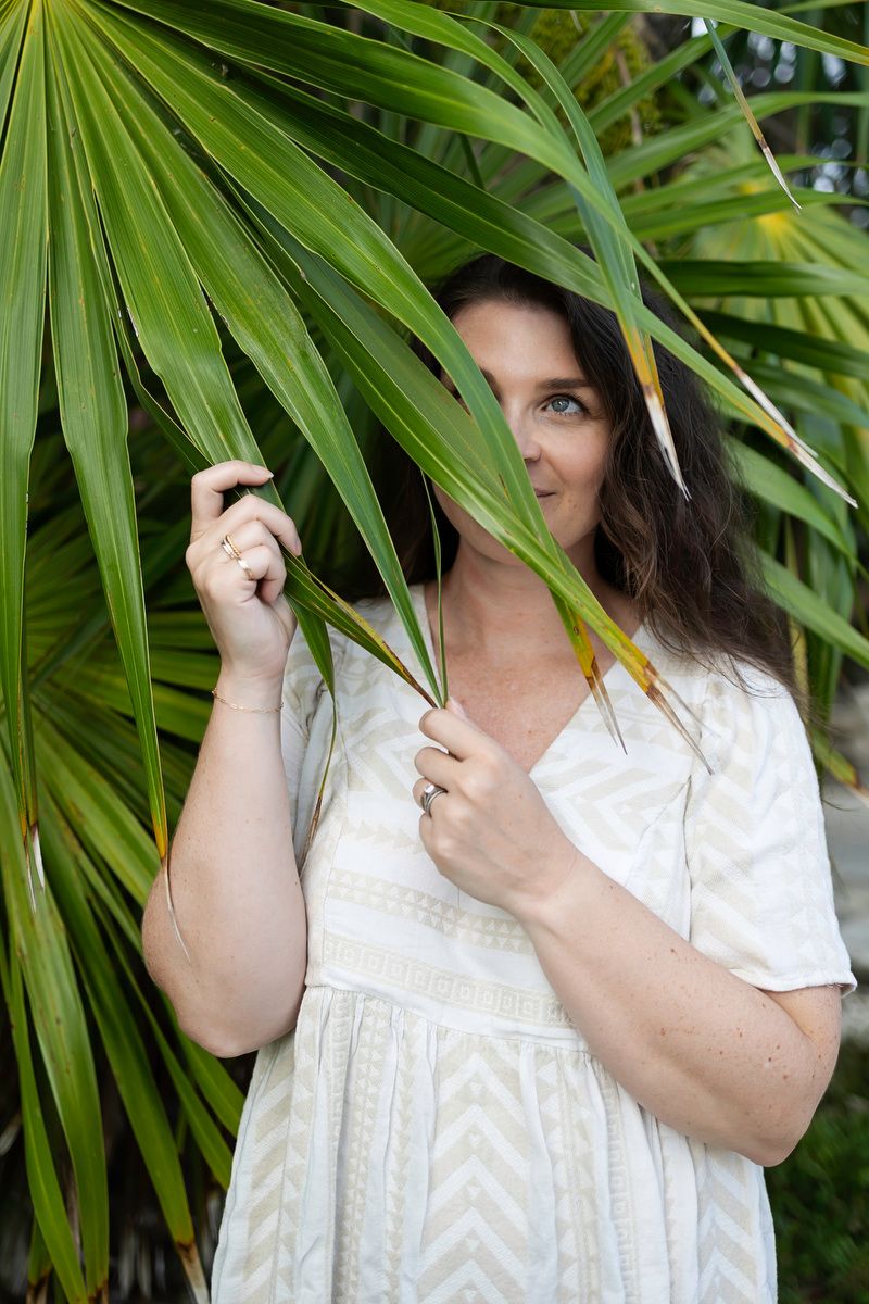 Woman in white dress partially hidden behind tropical palm leaves in a lush garden setting.