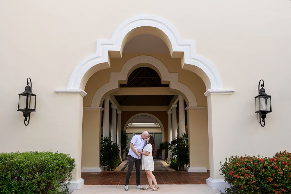 Couple embracing under elegant white arched entrance of a Mediterranean-style building with lantern wall sconces.