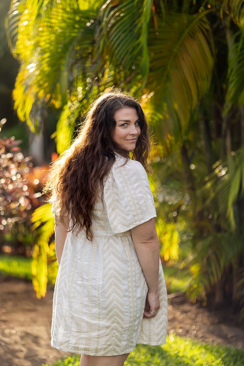 Young woman with long dark hair in white dress smiling over shoulder among tropical palm trees in golden sunlight.