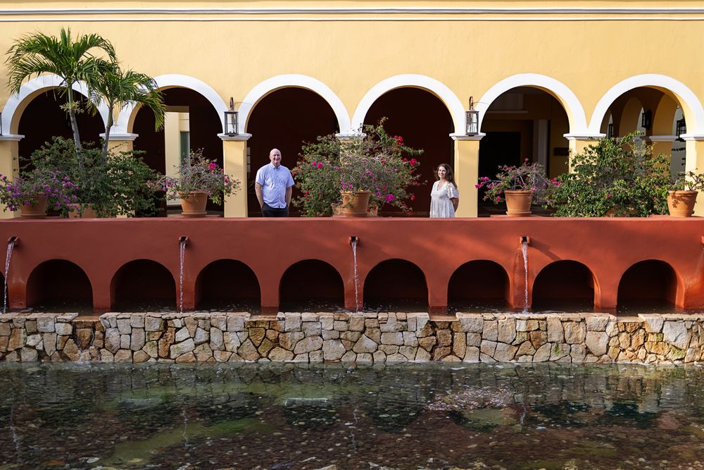 Two people stand on a terrace of a colorful Mexican hacienda with arches, bougainvillea, and a stone pool.