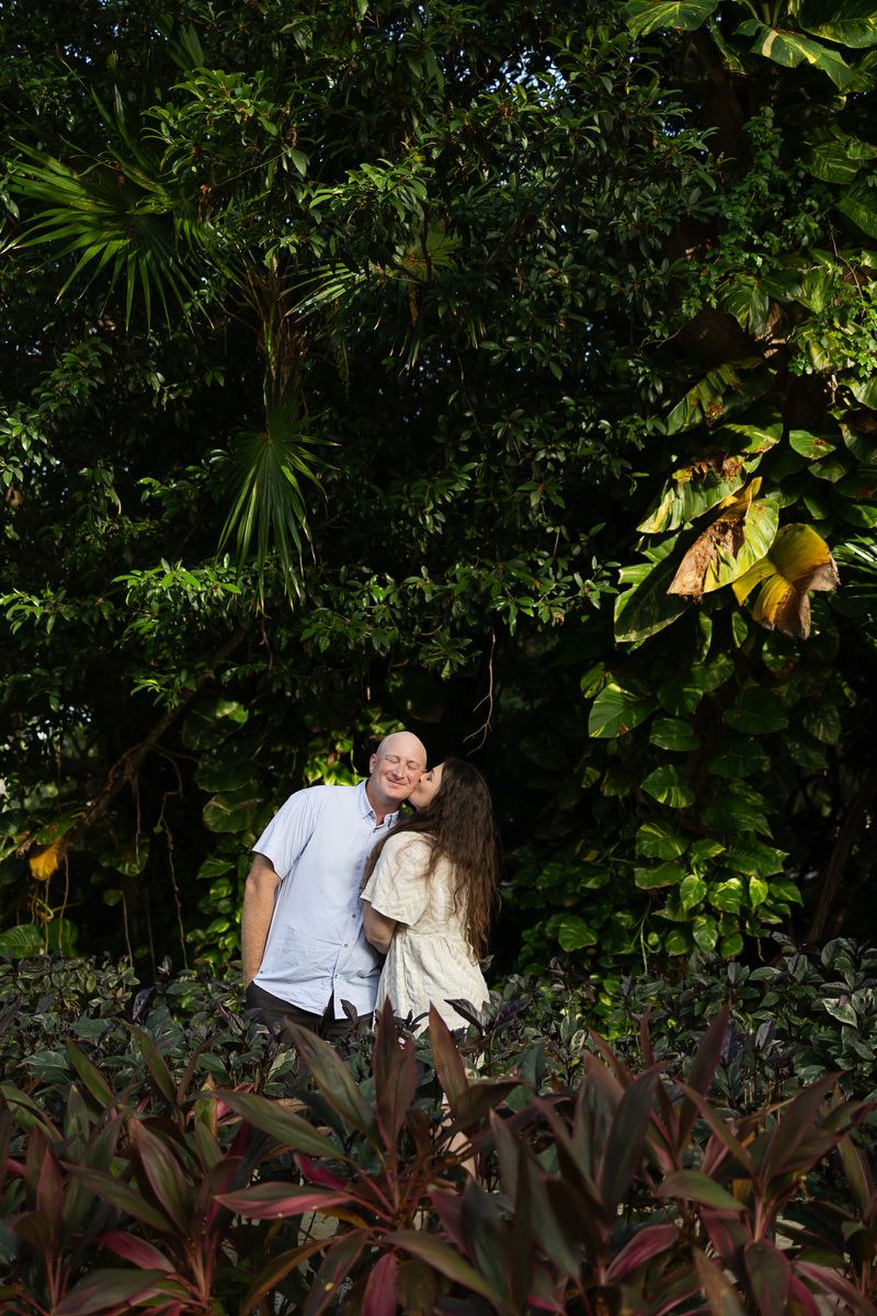 Couple sharing a kiss surrounded by lush tropical greenery and colorful foliage in a garden.