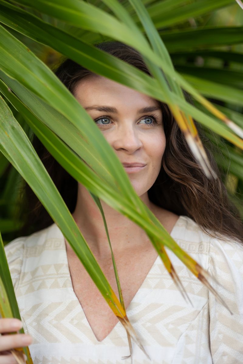 A woman with dark hair and blue eyes peers through lush green tropical palm leaves wearing a white patterned dress.