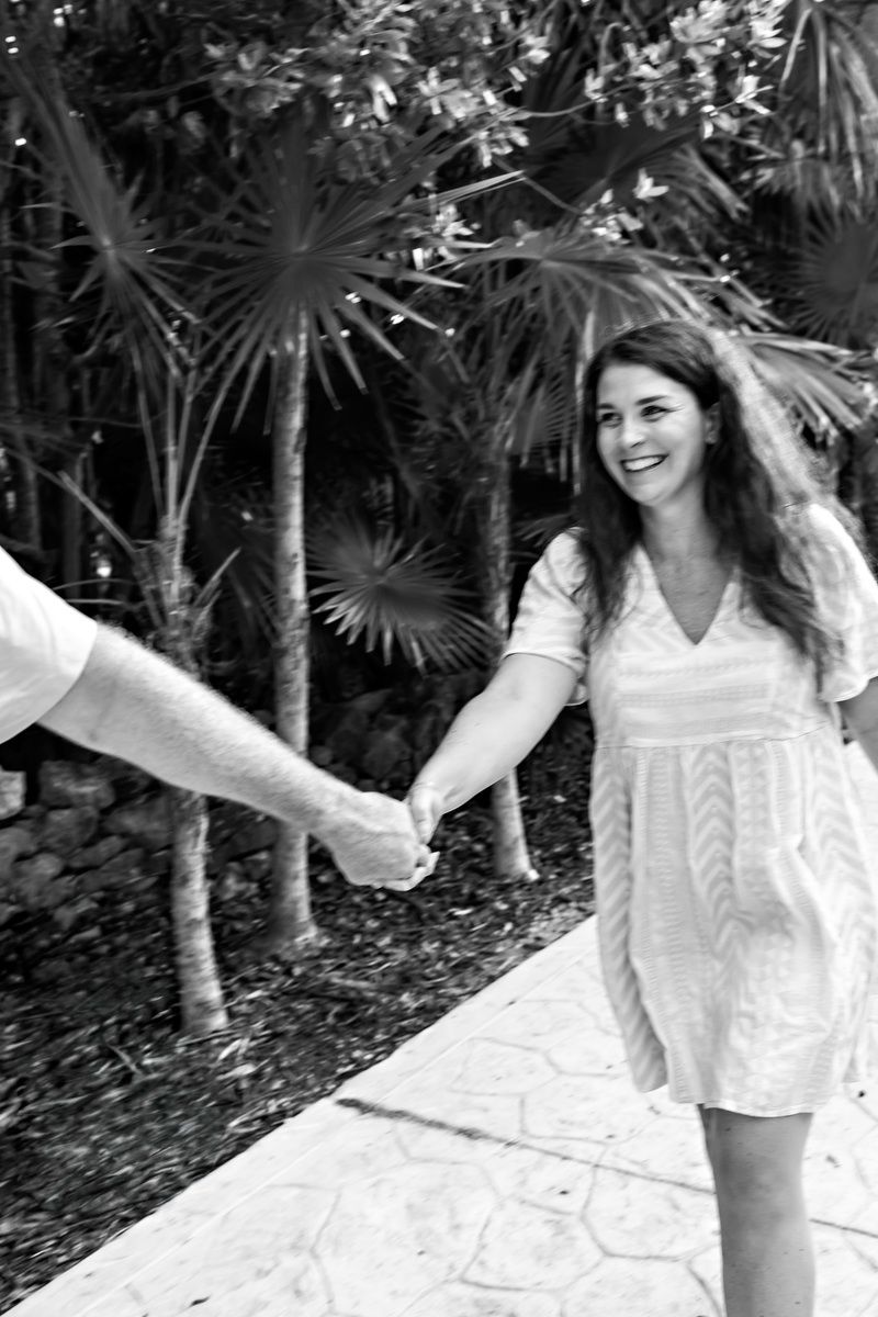 A smiling woman in a white dress holds hands with a man while walking on a tropical garden path in black and white.