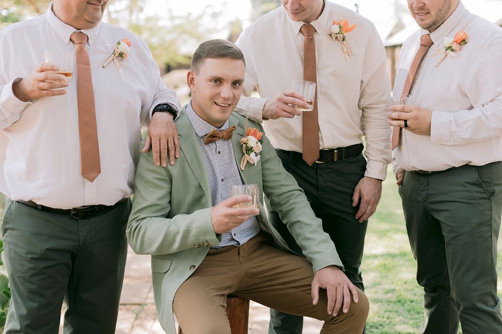 Groom in sage green suit celebrating with groomsmen wearing coral ties and floral boutonnieres.