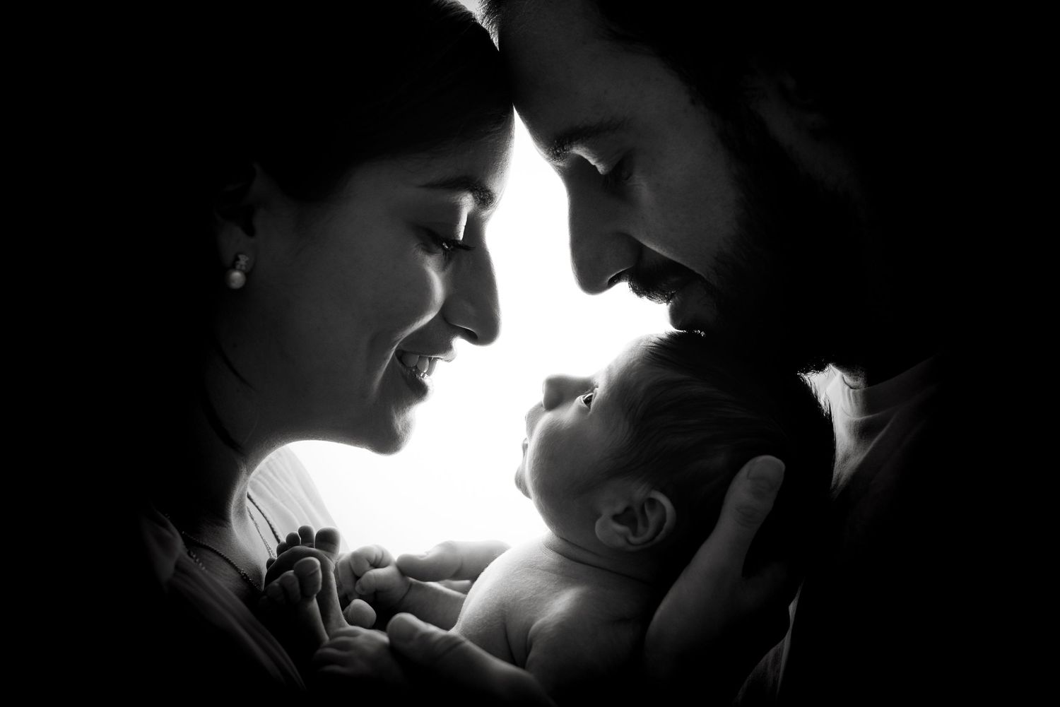 Black and white photo of parents lovingly gazing at their newborn baby held between them.