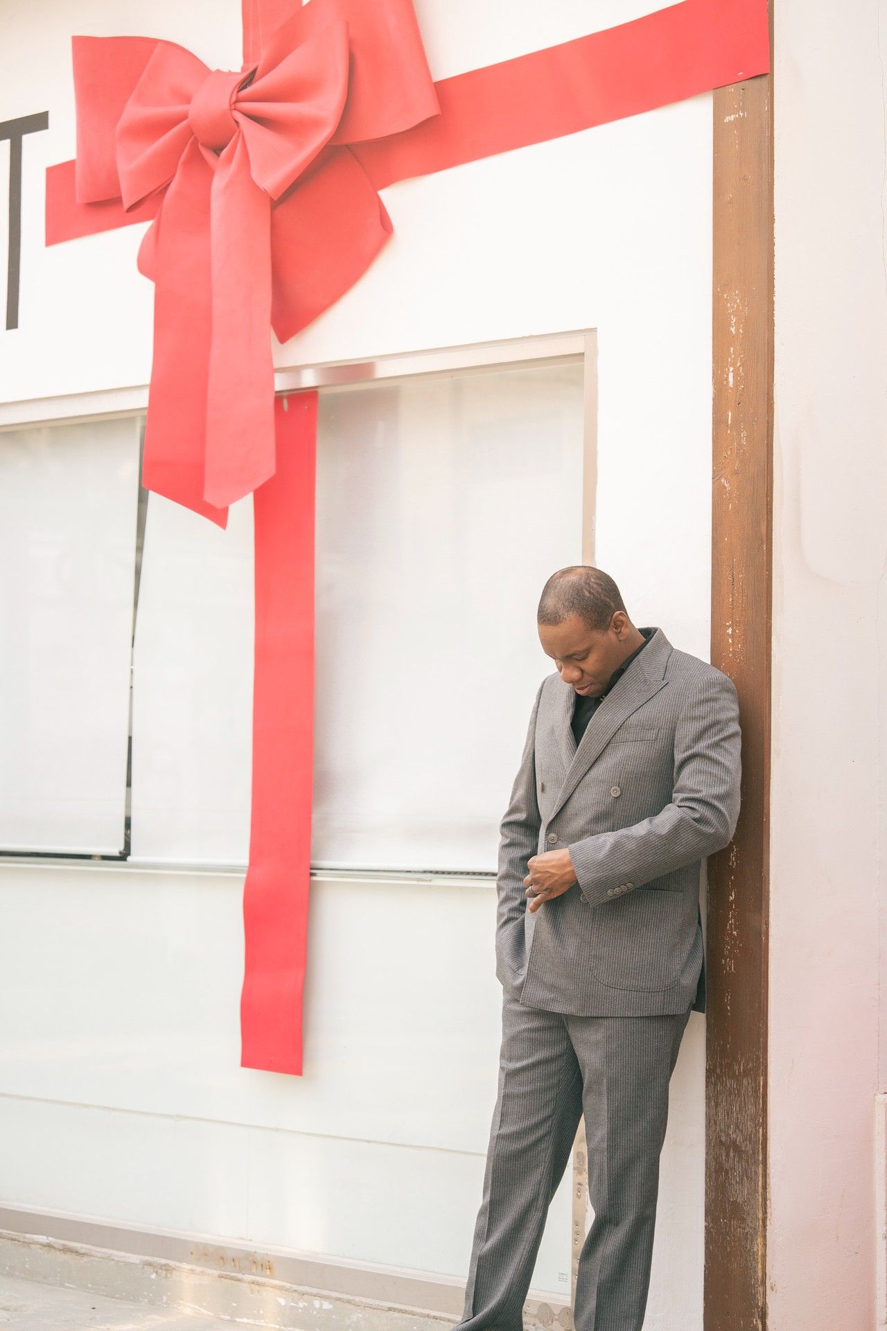 Man in gray suit leaning against wooden post near large decorative red bow and ribbon holiday display.