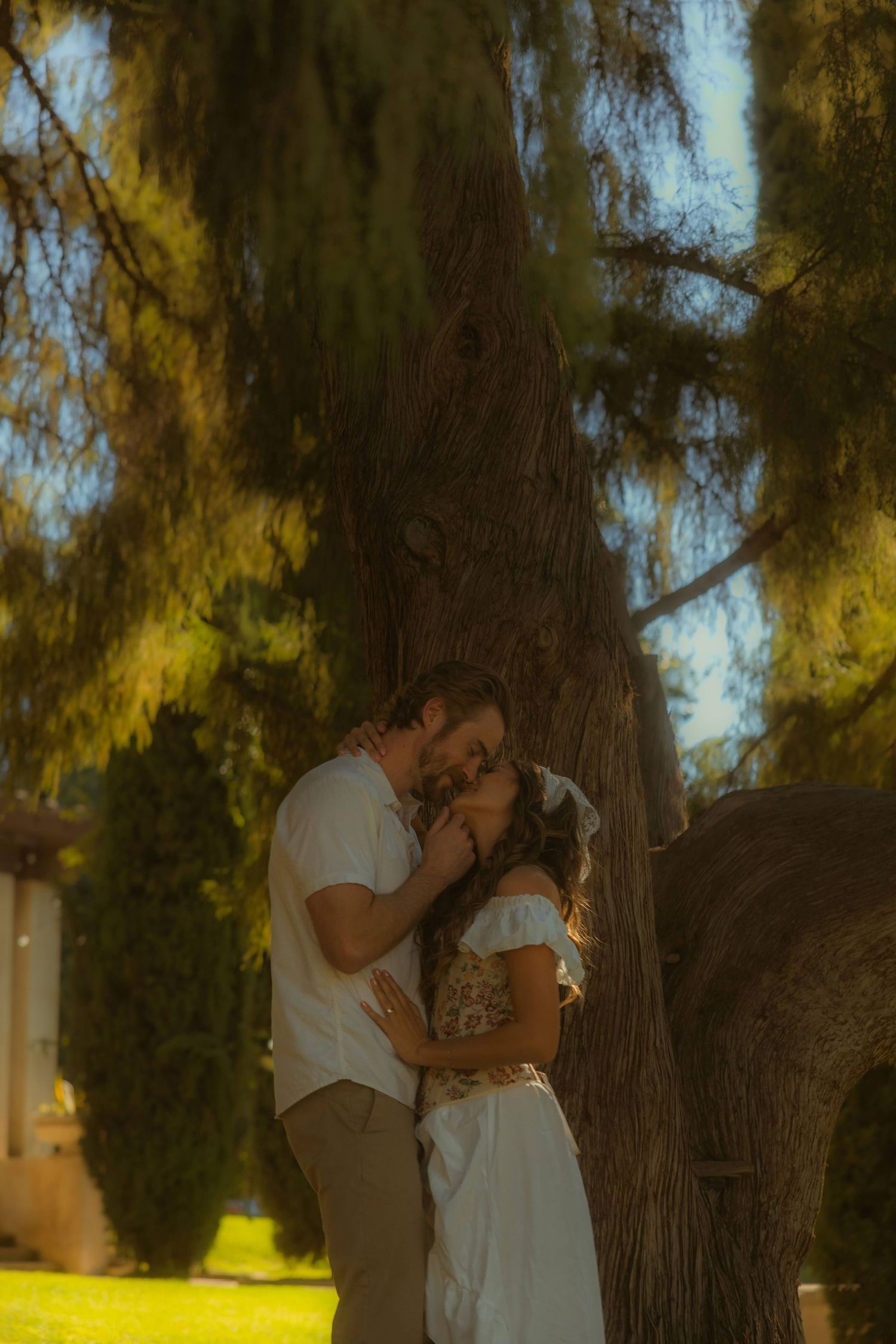 A couple shares a romantic kiss leaning against a large tree trunk on the grounds of the Kimberly Crest House wedding venue.