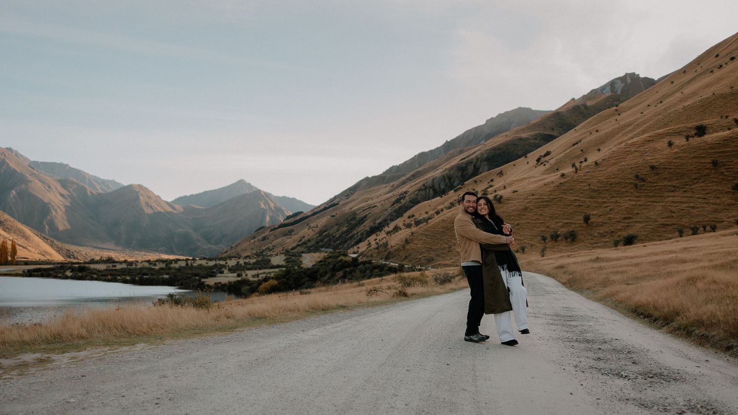 Couple dancing on a scenic gravel road surrounded by golden hills and a lake in New Zealand mountains.